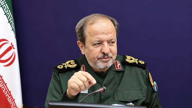 A senior Iranian military official in an olive-green IRGC uniform speaks at a podium with a microphone, with the Iranian flag visible in the background against a blue backdrop.