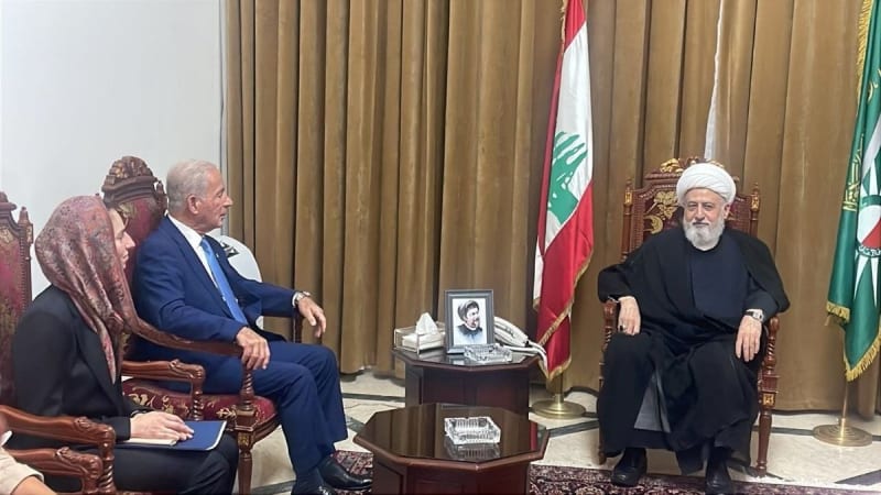 A formal diplomatic meeting in Lebanon showing a Shia cleric in black robes and white turban seated across from a man in a blue suit, with a woman holding documents nearby. Lebanese and green flags are visible in the background, along with a framed photograph on the table.