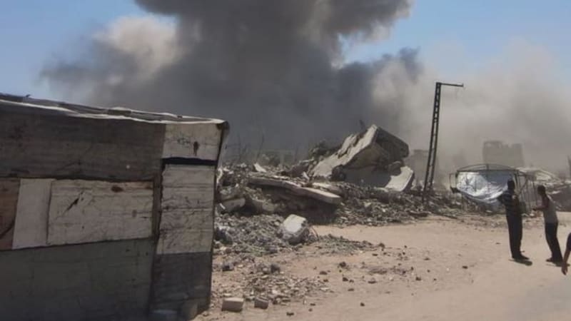 A large plume of smoke rises into a clear sky above a heavily damaged urban area with rubble and destroyed buildings, with a person standing nearby observing the aftermath of what appears to be an airstrike or explosion.