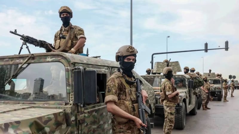 Armed soldiers in camouflage uniforms with face coverings stand beside and atop a convoy of military vehicles equipped with mounted machine guns on an urban road, likely in a Middle Eastern or North African setting.