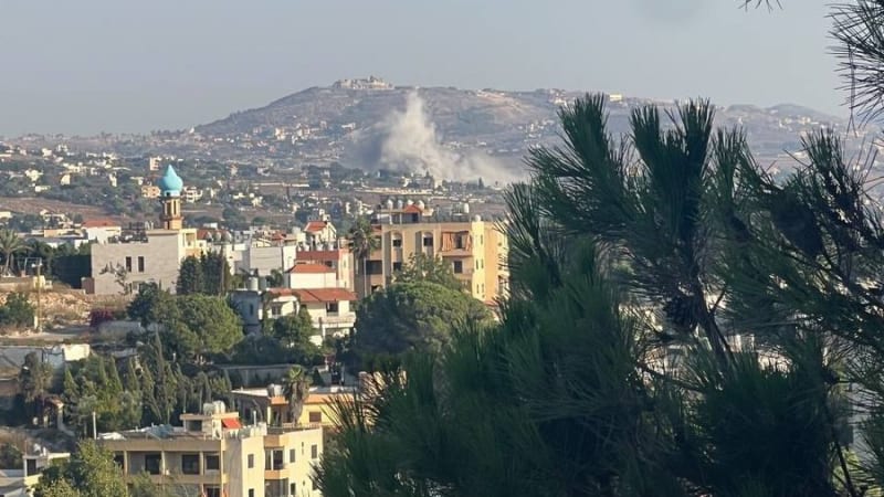 Smoke rising in the distance over a Lebanese town with residential buildings, a mosque minaret, and pine trees in the foreground, consistent with an airstrike or shelling in southern Lebanon.