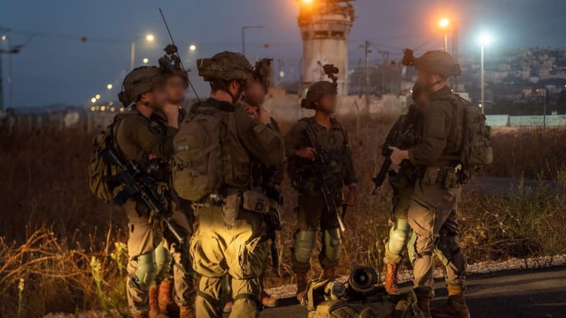 A group of Israeli Defense Forces (IDF) soldiers with blurred faces gather at dusk near a military watchtower, equipped with tactical gear, weapons, and night vision devices, likely in the West Bank.