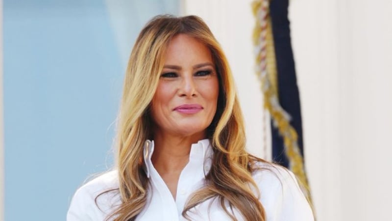 A woman with long blonde-highlighted hair wearing a white blouse, photographed at what appears to be an official or formal setting with a flag visible in the background.