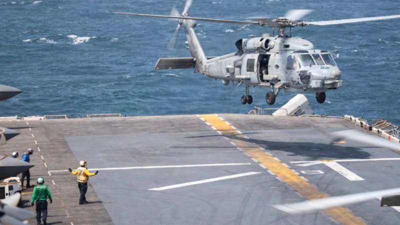 A U.S. Navy MH-60 Seahawk helicopter (number 704) prepares to land on the flight deck of a warship at sea, with deck crew in colored jerseys directing operations.