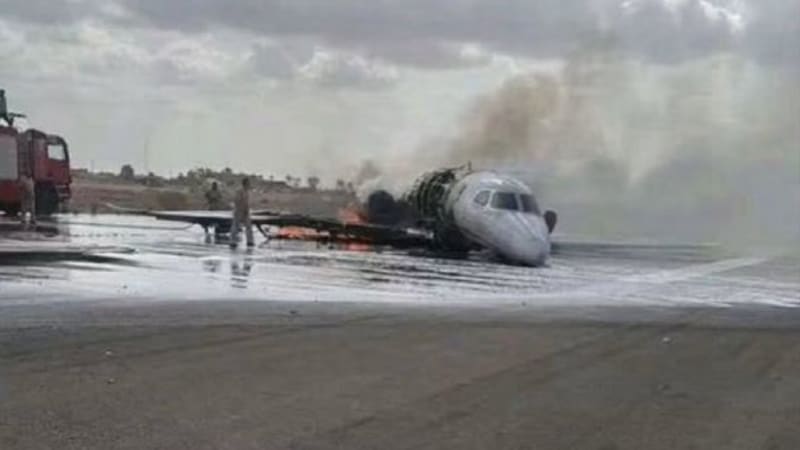 A damaged aircraft on fire on an airport runway with smoke rising, firefighting foam visible on the tarmac, and a fire truck responding to the incident.