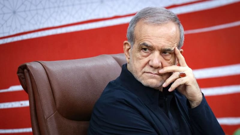 An older man in a dark shirt sits in a brown leather chair with his hand resting against his face, wearing an orange agate ring, with a red and white backdrop behind him.