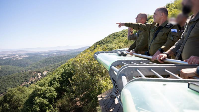 Israeli Defense Forces (IDF) officers stand at an elevated observation post overlooking green, hilly terrain near a border area, with one officer pointing into the distance while others observe with binoculars nearby.