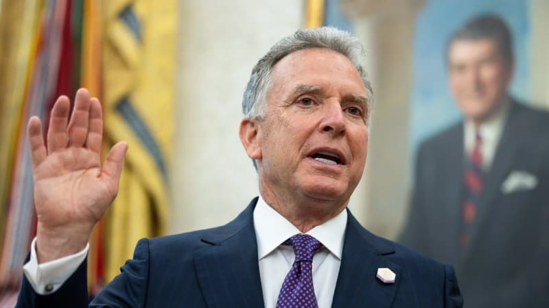 A man in a dark pinstripe suit with a polka-dot tie raises his right hand as if taking an oath, with a portrait painting visible in the background.
