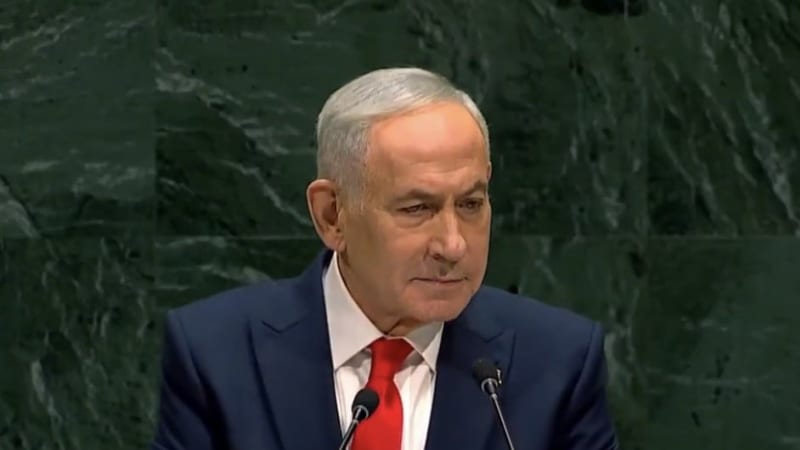 A man in a blue suit with a red tie speaks at a podium with microphones, standing before the distinctive green marble backdrop of the United Nations General Assembly hall.