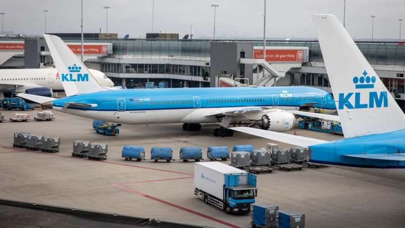 KLM Royal Dutch Airlines aircraft parked at a gate at Amsterdam Schiphol Airport, with ground service equipment, cargo containers, and a KLM catering truck on the tarmac.