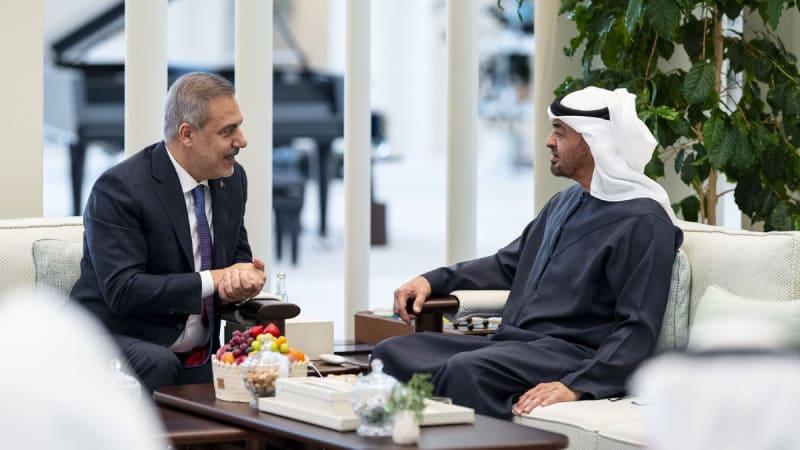 Two officials engaged in a diplomatic meeting, one wearing a Western business suit and the other in traditional Gulf Arab attire including a white headscarf and dark robe, seated in a modern reception area with a fruit platter on the table between them.