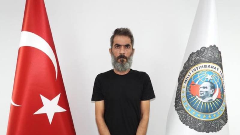 A handcuffed man in a black shirt standing between a Turkish national flag and a flag bearing the emblem of Turkey's National Intelligence Organization (MİT), in what appears to be an official detention or arrest photo.