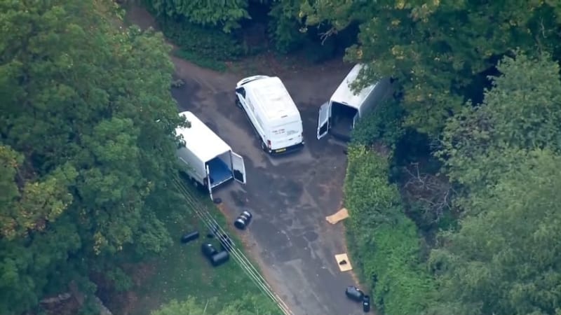 Aerial view of three white vans parked in a wooded clearing with their doors open, surrounded by dense green trees, with some dark objects on the ground nearby, suggesting a law enforcement or investigative scene.