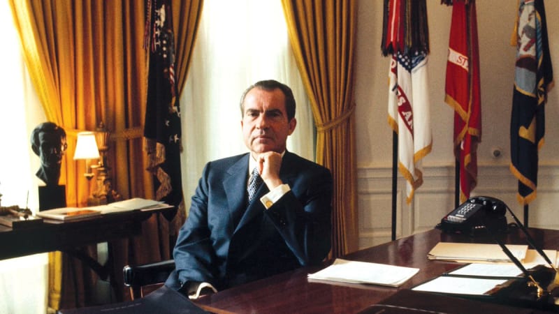 A man seated at a desk in the Oval Office of the White House, with gold curtains, American flags, and a telephone visible in the background.