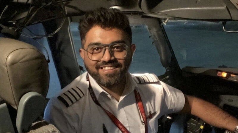A smiling man wearing a pilot's uniform with epaulettes and glasses sits in an aircraft cockpit, with instruments and controls visible in the background.