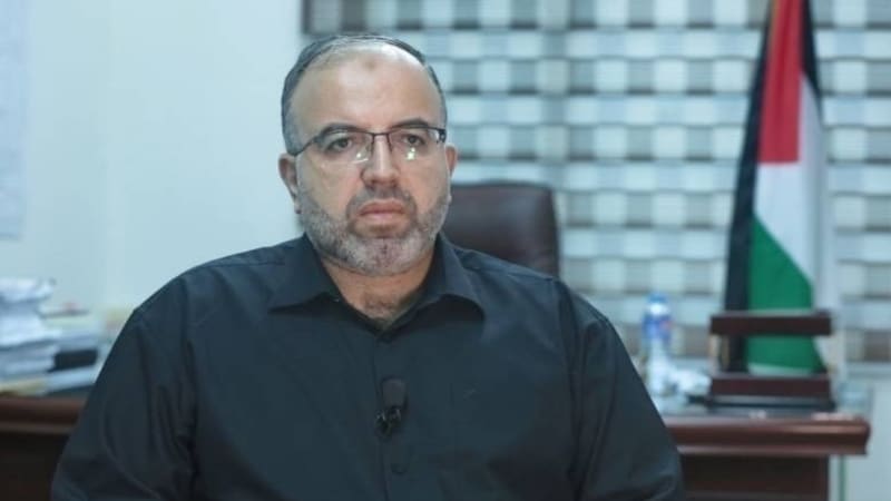 A man wearing glasses and a dark shirt sits in an office setting with a Palestinian flag visible in the background.