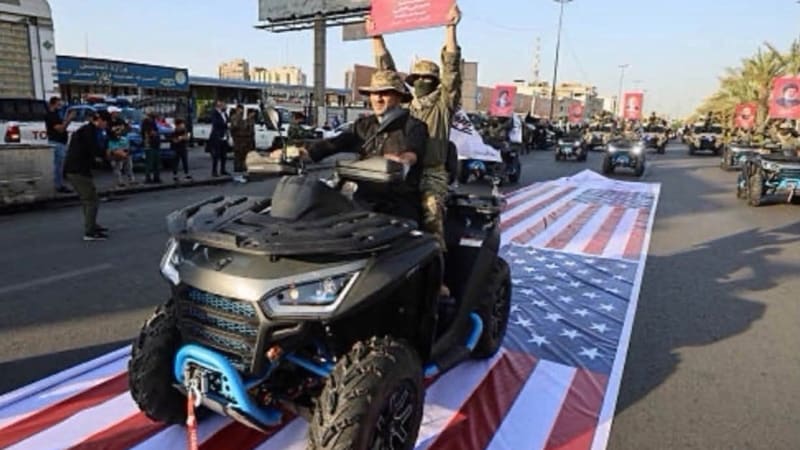 Militants on ATVs drive over a large American flag laid on the ground during a parade, with a passenger holding up a portrait of a religious leader. Additional vehicles with similar portraits are visible in the convoy on an urban street.