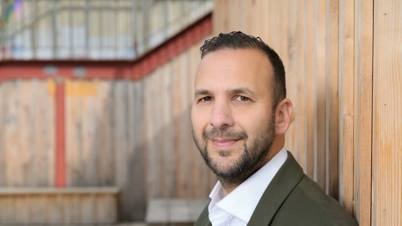 Portrait of a bearded man wearing a white shirt and olive green blazer, leaning against a wooden wall with a wooden deck and structure in the background.