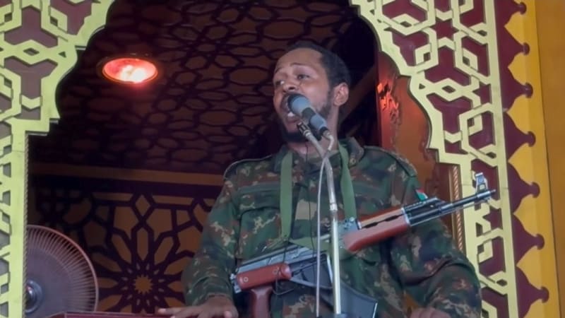 A man in military camouflage uniform carrying an AK-style rifle speaks into a microphone from what appears to be a mosque pulpit (minbar) with ornate Islamic geometric patterns in the background.