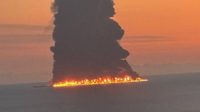 A massive fire burns along a wide front, producing a towering column of thick black smoke rising into an orange sunset sky, viewed from a distance over residential apartment buildings.