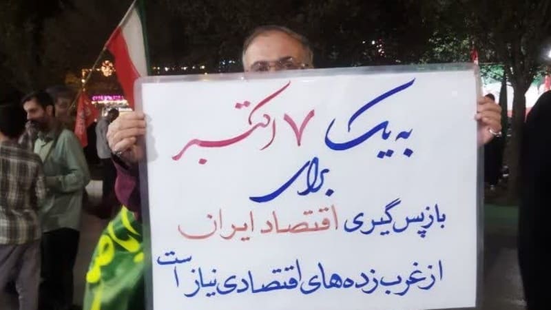A man at a nighttime rally in Iran holds a handwritten sign in Persian calling for reclaiming Iran's economy from Western-influenced economic figures, with Iranian flags visible in the background.