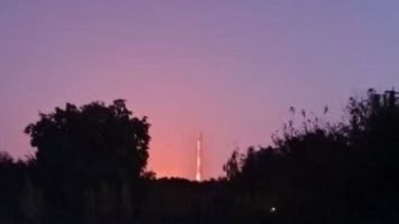 A rocket launch seen from a distance at dusk or dawn, with a bright exhaust trail visible above a treeline against a purple-pink sky.