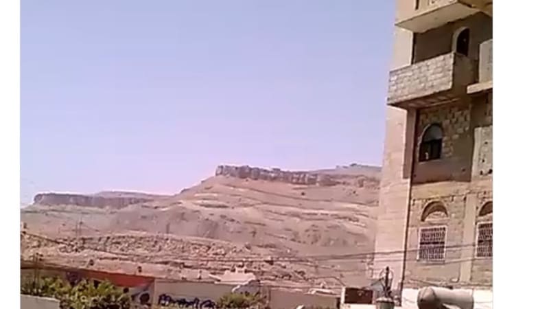 Photo taken from under a covered balcony or archway showing arid mountainous terrain and a stone building in what is described as southwest of Sanaa, Yemen, with overlaid text referencing heavy bombing and possible secondary explosions.