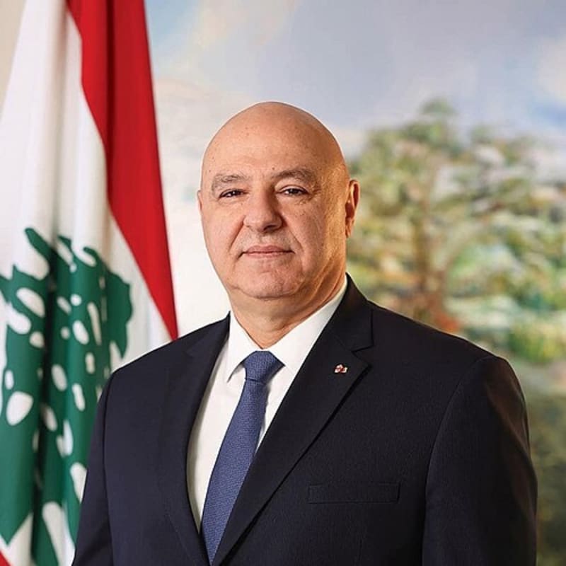 Official portrait of a bald man in a dark navy suit and blue tie, standing in front of the Lebanese flag and a painting of cedar trees.