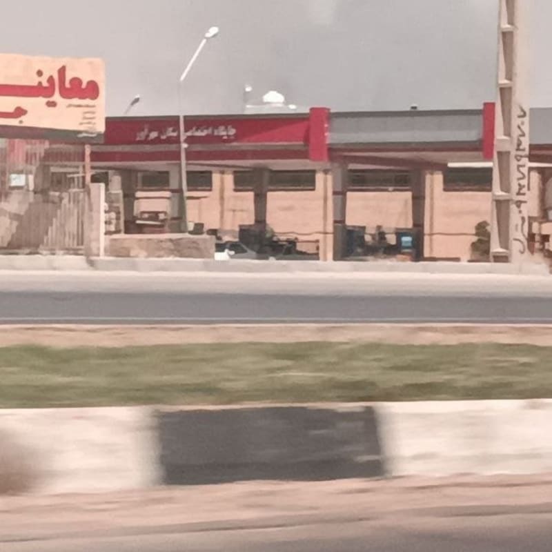 A roadside gas station or vehicle inspection center with Arabic/Persian script signage, photographed from across a highway under hazy or smoky skies with dark smoke visible in the background.