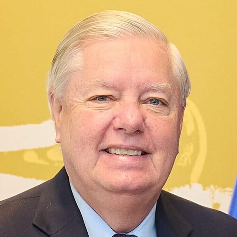 Portrait of an older man with white hair wearing a dark suit, light blue shirt, and patterned tie with a small flag pin on his lapel, against a yellow background.