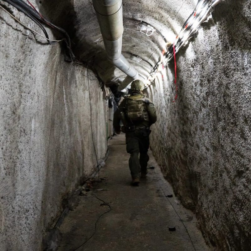A soldier in full tactical gear walks through a narrow underground tunnel with concrete walls, visible piping, and wiring along the ceiling, consistent with military tunnel operations in Gaza.