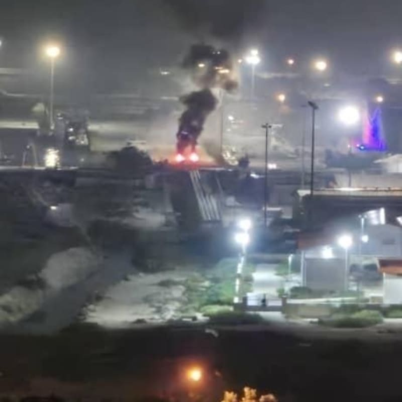 Nighttime fire at what appears to be a port or industrial waterfront area, with thick black smoke rising from flames visible among lit infrastructure and docked vessels.