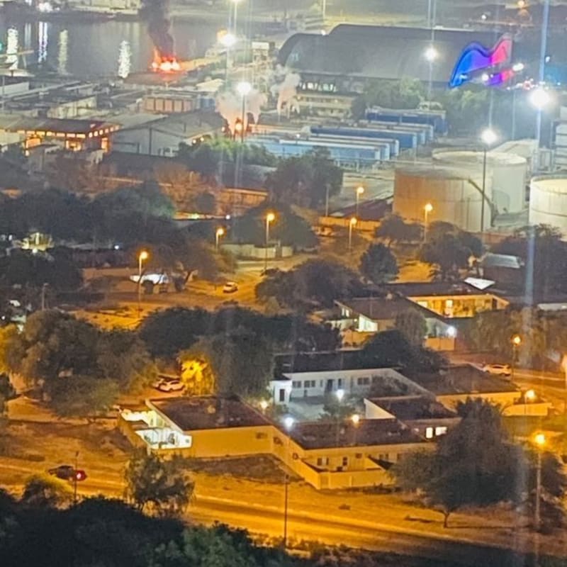 Nighttime elevated view of a port or industrial waterfront area with a large fire producing thick black smoke, visible among harbor infrastructure, storage tanks, and illuminated buildings.