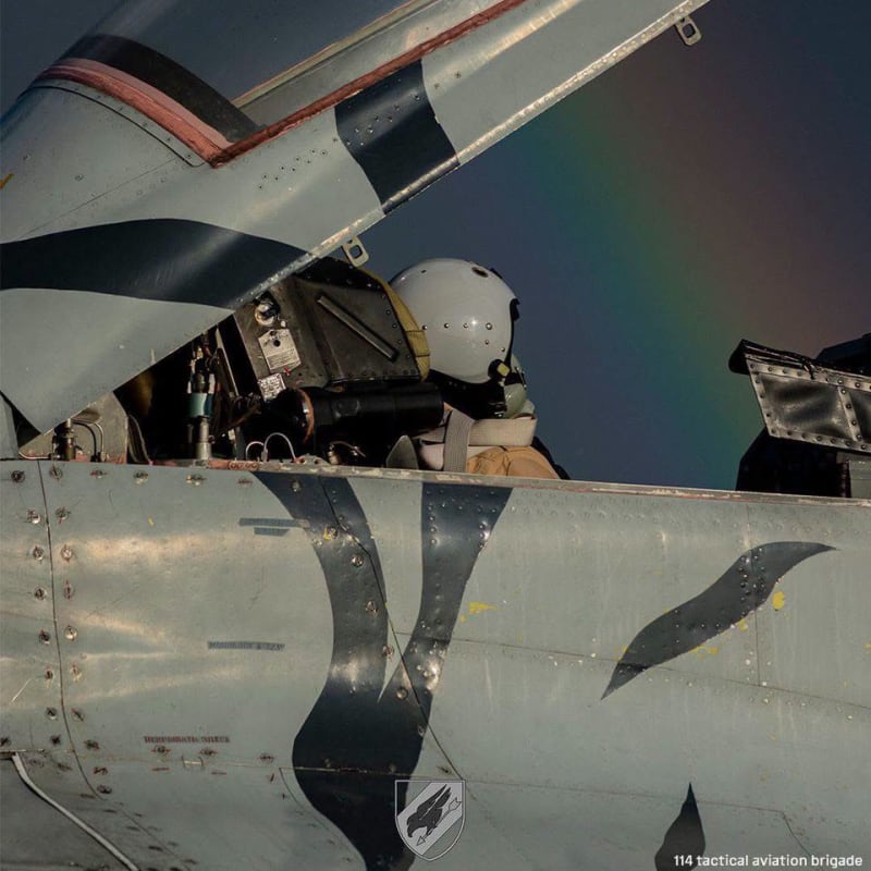 A pilot wearing a white helmet sits in the open cockpit of a military fighter jet with digital camouflage paint scheme, credited to Ukraine's 114th Tactical Aviation Brigade.