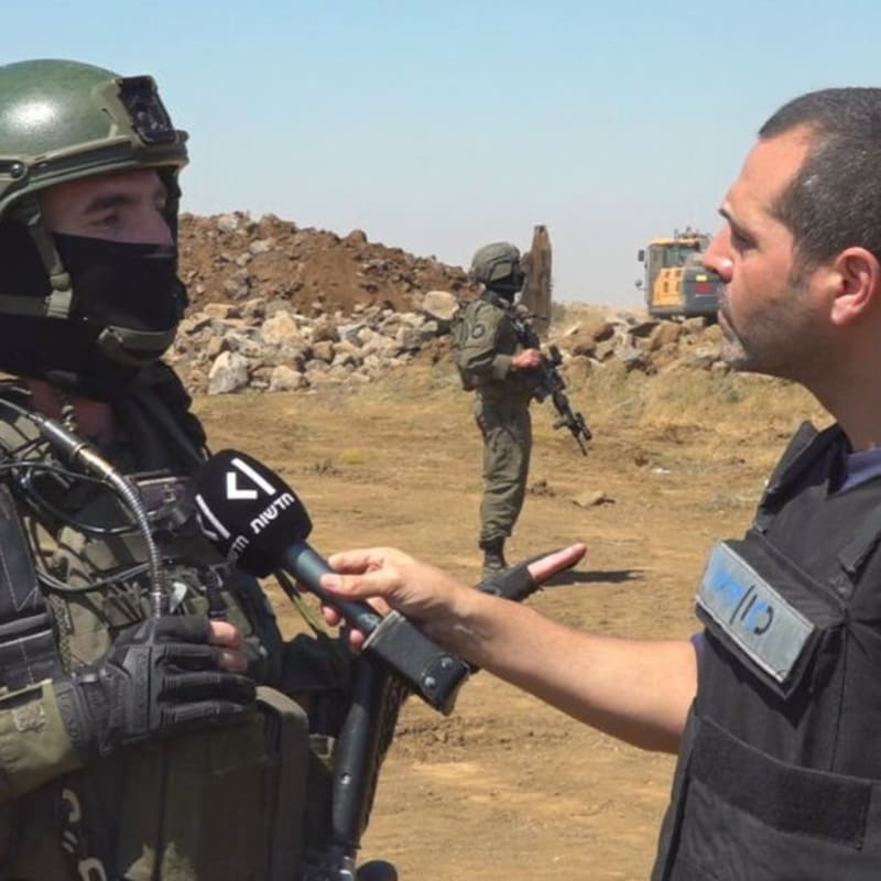 A journalist wearing a bulletproof vest interviews a masked Israeli soldier in full combat gear at what appears to be a military operational area with earth mounds and heavy machinery in the background. The microphone bears Hebrew-language news branding.