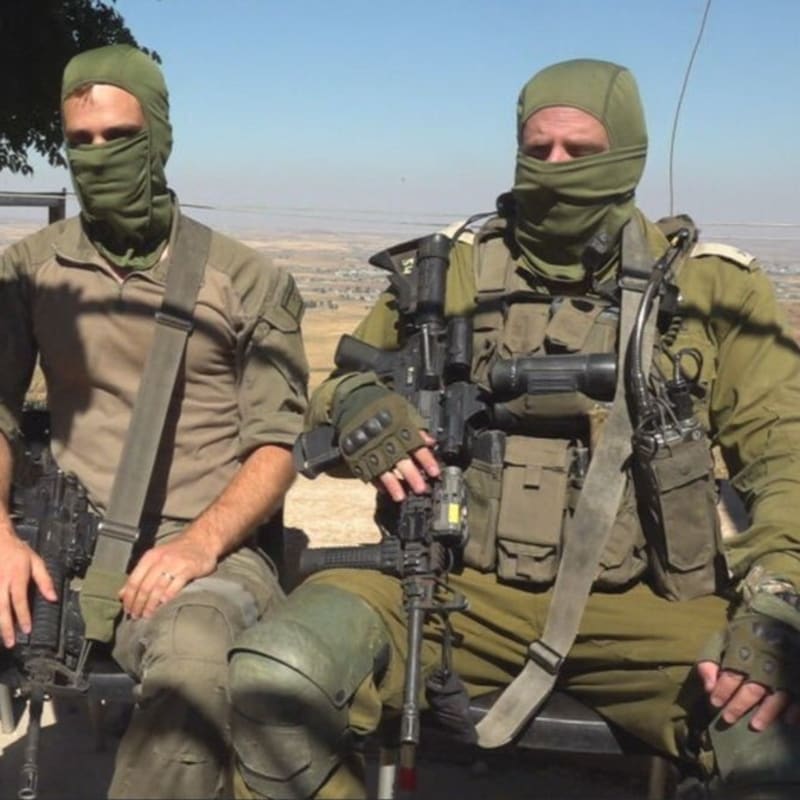 Three soldiers in olive-green military uniforms and face-covering balaclavas sit together holding assault rifles, with an arid hilly landscape visible in the background.
