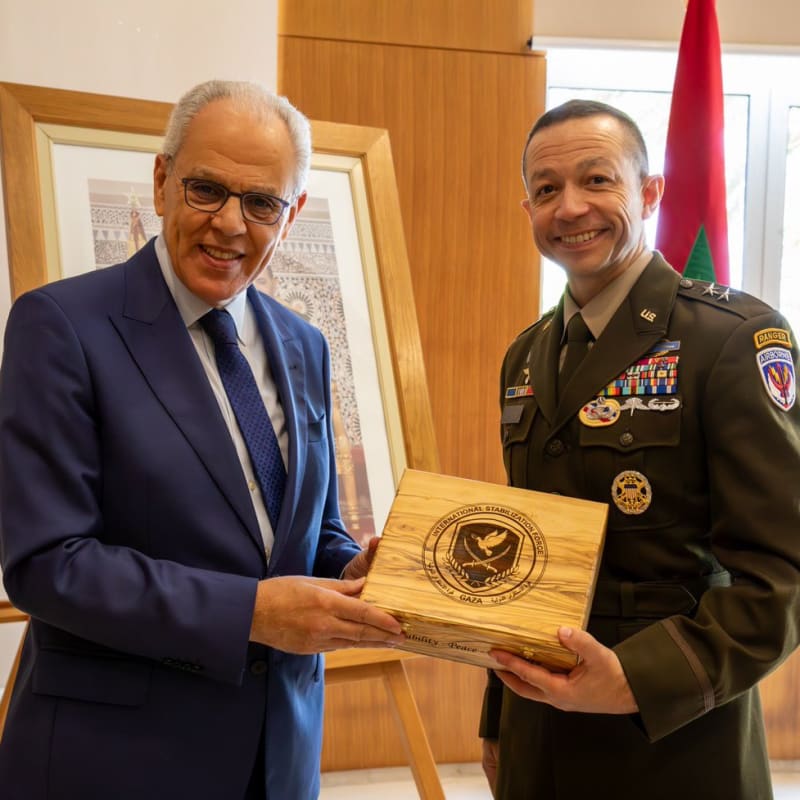 A civilian in a blue suit and a U.S. Army officer wearing Ranger and Airborne tabs pose together holding a wooden box engraved with the International Stabilization Force Gaza emblem and the words 'Stability Peace,' with a Moroccan flag visible in the background.