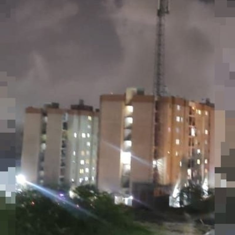 Nighttime photo of a multi-story residential apartment building with a telecommunications tower on top, set against a cloudy sky with some illuminated windows visible.