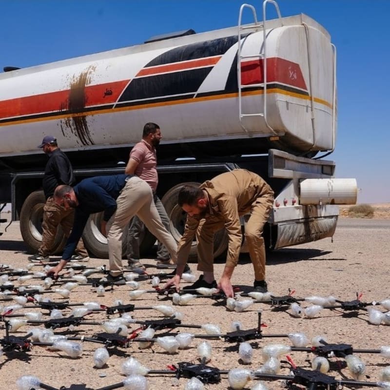 Several men in a desert environment unload and arrange dozens of small black quadcopter drones and plastic-wrapped packages from a fuel tanker truck, dated 2026/07/16 in what appears to be a Middle Eastern setting.