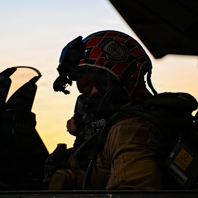 A military fighter pilot wearing a red checkered helmet sits in an open cockpit silhouetted against a sunset sky.