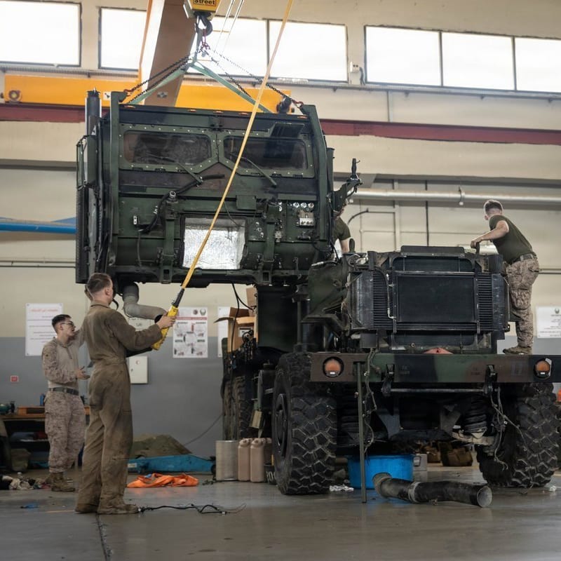 U.S. Marines perform maintenance on a military tactical vehicle inside a maintenance bay, using an overhead crane to lift the armored cab from the chassis.