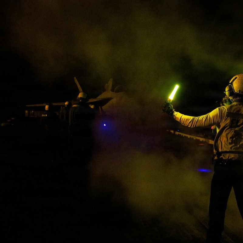 A flight deck crew member wearing a helmet and protective gear directs aircraft at night on an aircraft carrier, holding a glowing yellow wand amid steam or exhaust, with a military jet visible in the background.