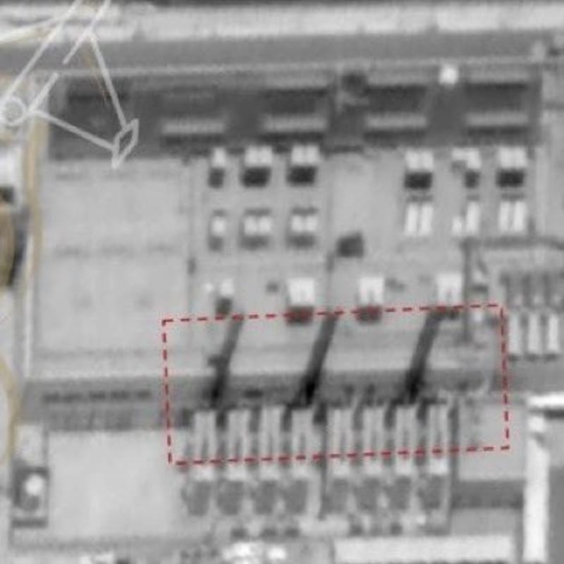 Grayscale satellite imagery showing an overhead view of an industrial or military facility with a red dashed rectangle highlighting a specific section of the building complex.