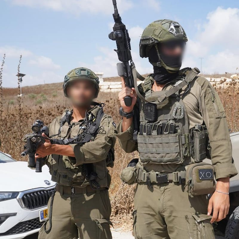 Two Israeli soldiers with blurred faces wearing olive-green uniforms, tactical vests, helmets, and carrying assault rifles, standing near military and civilian vehicles in an arid landscape.