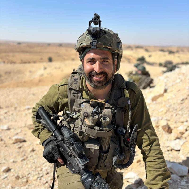 A smiling soldier in IDF olive-green uniform and tactical gear, including a combat helmet with NVG mount, body armor, radio equipment, and an assault rifle, posing in an arid desert landscape with other soldiers visible in the background.