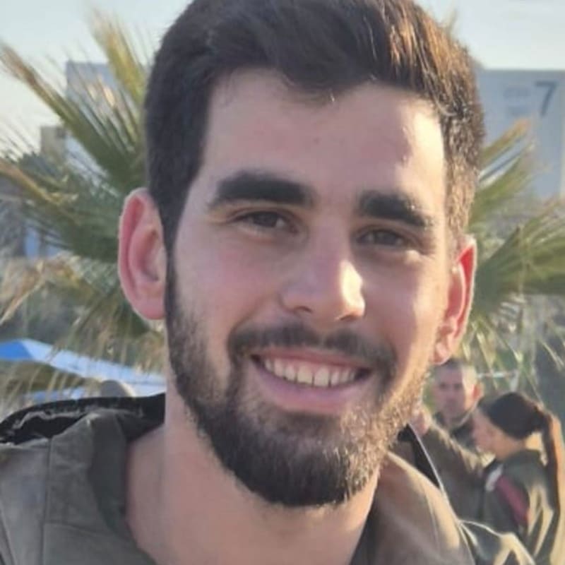 A young bearded man smiling at the camera while wearing an olive-green military uniform and tactical vest, with palm trees and other uniformed personnel visible in the background.