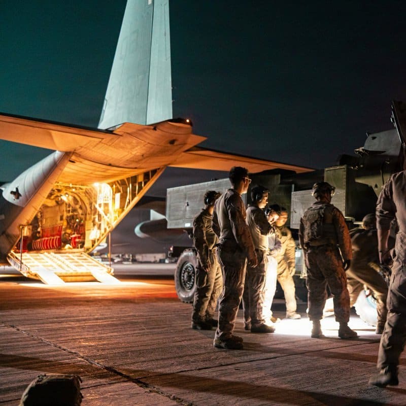 Military personnel stand on a tarmac at night preparing to load or unload a HIMARS rocket launcher system from the open rear cargo ramp of a C-130 transport aircraft.