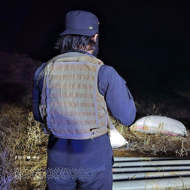 A man wearing a tactical vest and bucket hat stands at night with his back to the camera, overlooking what appears to be rocket or missile tubes and sandbags on the ground in an arid area.