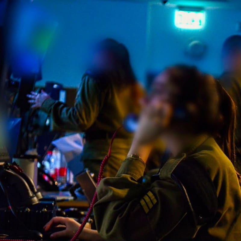 Military personnel in olive-green uniforms working at computer stations in a dimly lit command or operations center, with faces deliberately blurred for security. The lighting is predominantly blue, suggesting screen-lit conditions typical of a military operations room.