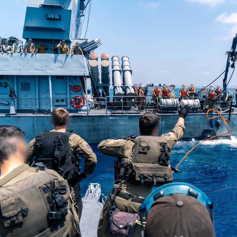 Naval personnel throwing a line between two warships during an underway replenishment or alongside transfer operation at sea, with sailors in tactical gear and life vests visible on both vessels. The warship appears to be an Israeli Navy Sa'ar-class corvette equipped with missile launchers.